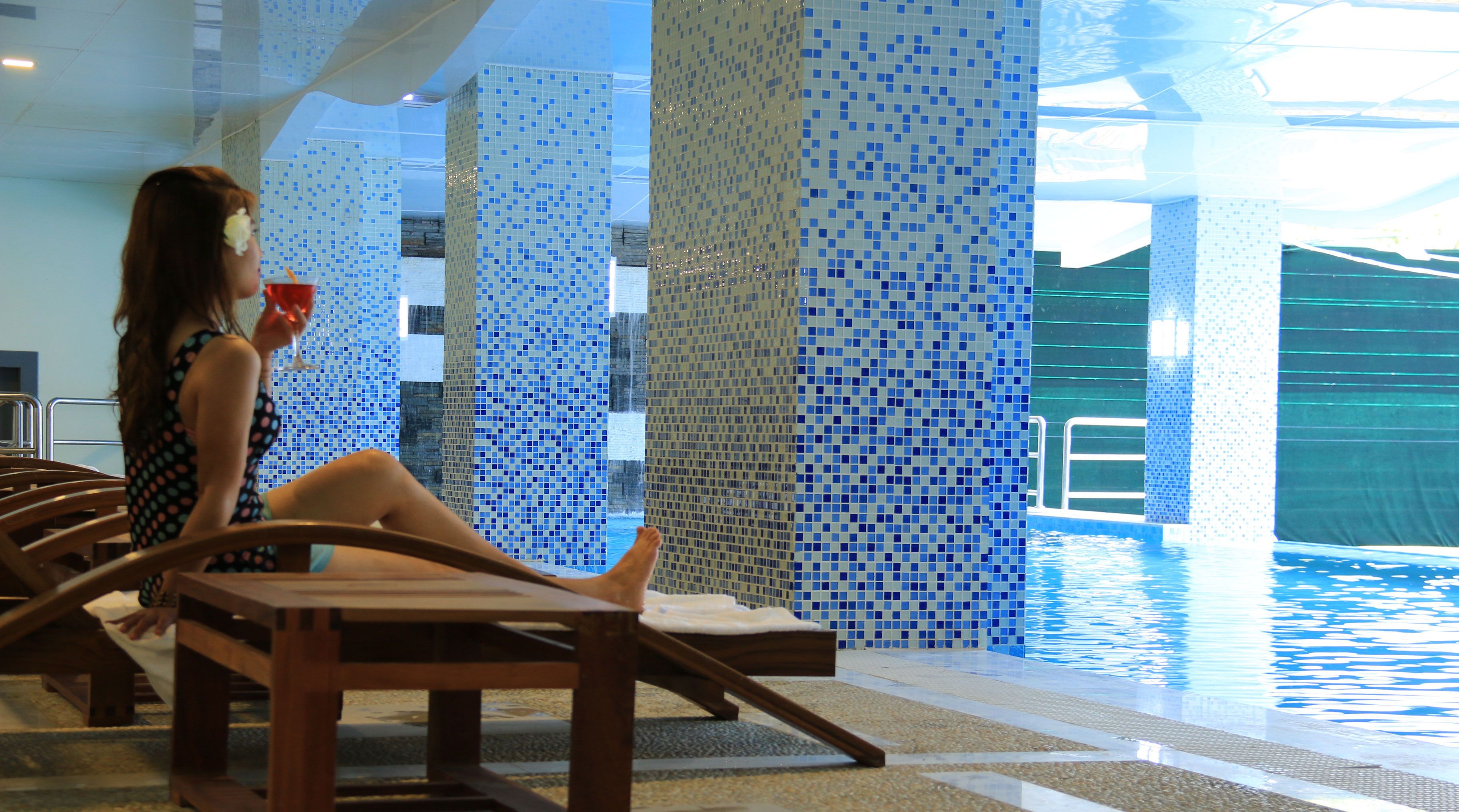 A person relaxing by a poolside lounge chair with a drink, surrounded by a tiled pool area with a view of the water.