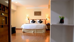 A hotel room with a large bed, two bedside lamps, a wooden floor, a wall-mounted artwork, and a small potted plant on a shelf. - Eastern Palace Hotel (Mandalay)