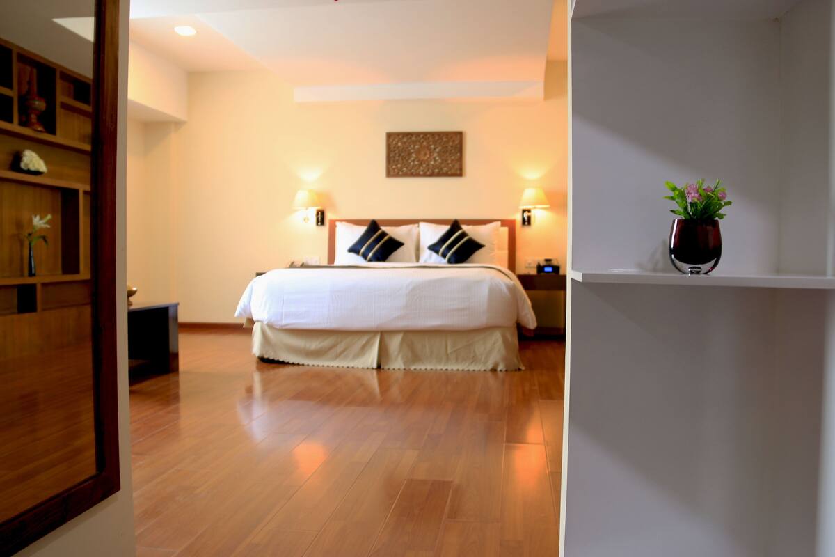 A hotel room with a large bed, two bedside lamps, a wooden floor, a wall-mounted artwork, and a small potted plant on a shelf.