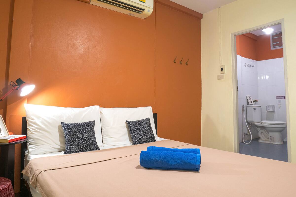 A hotel room with a bed, a desk with a lamp, an air conditioner, and a bathroom visible through an open door.