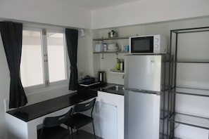 Una cocina compacta con cubierta negra, armarios blancos, microondas, refrigerador y una ventana con cortinas negras.
