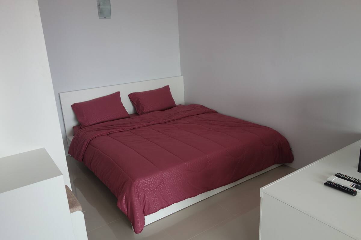 Un lit bien fait, avec une couverture rouge et des oreillers, une table de chevet blanche sur laquelle reposent deux télécommandes, et un mur blanc.