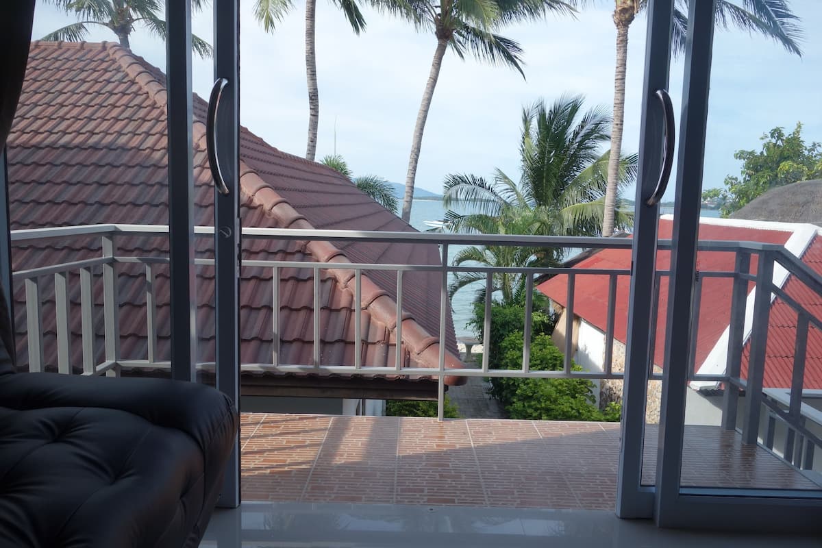 Un balcon avec une porte vitrée, un canapé en cuir noir et une vue sur un immeuble aux toits rouges et des palmiers.
