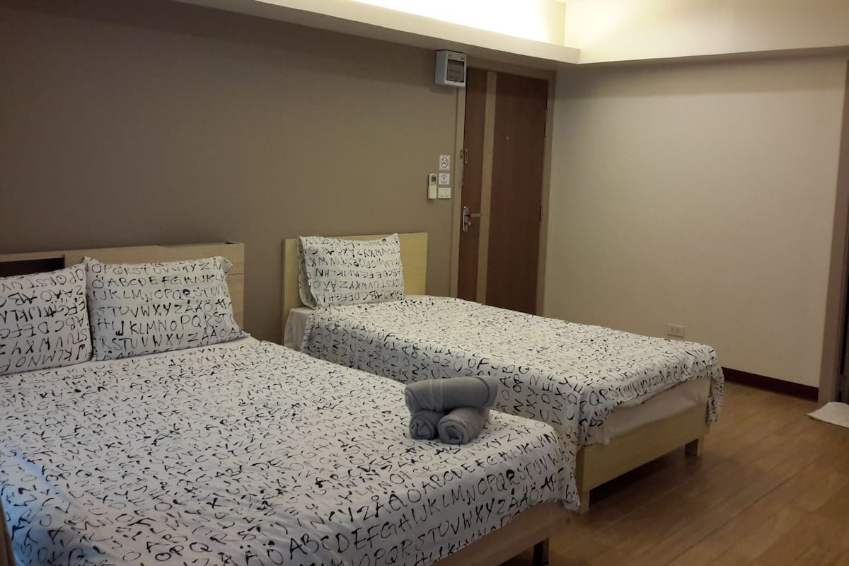 A hotel room with two beds, each with a white bedspread featuring black letters. There is a small table with a rolled-up towel on it.