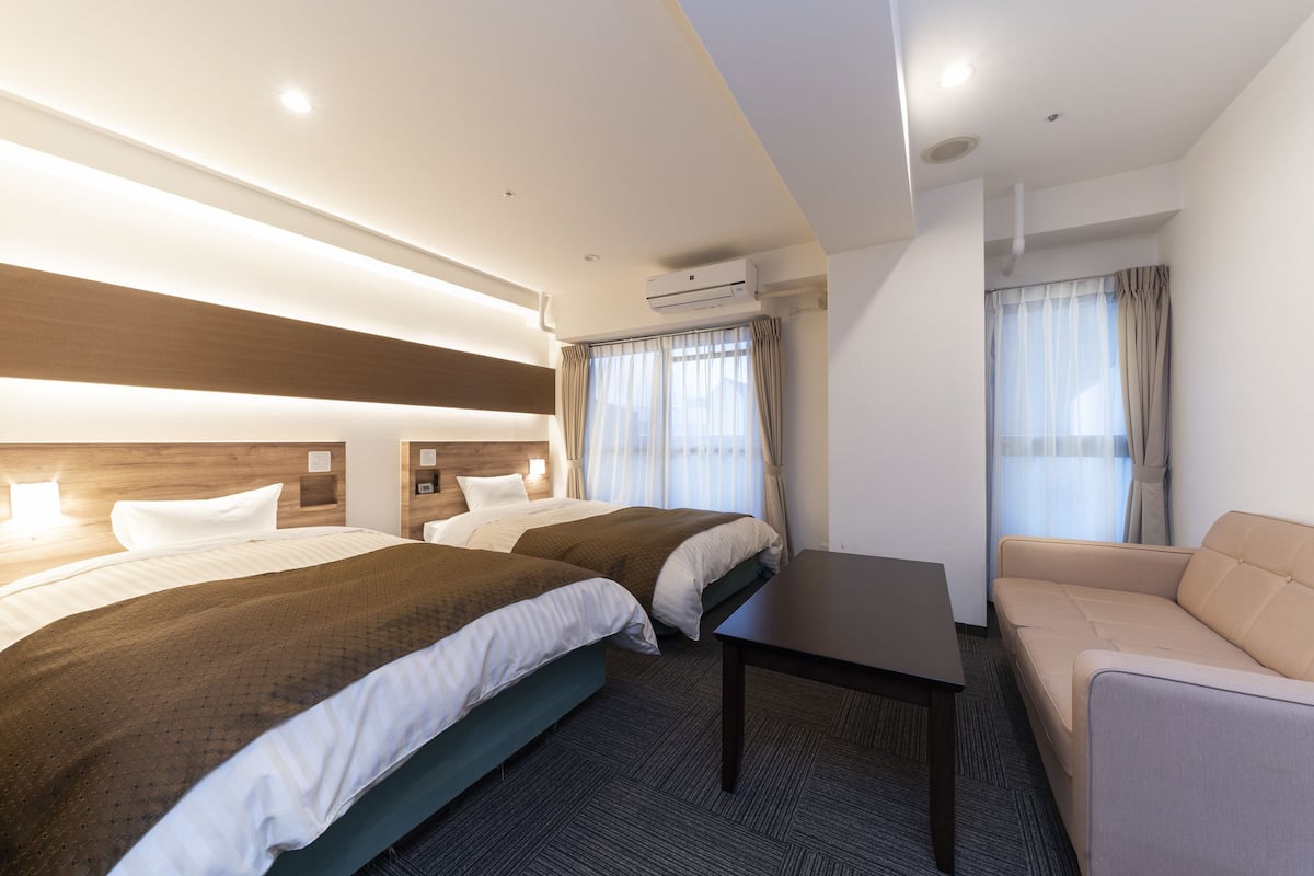 A hotel room with two beds, a sofa, a table, and an air conditioning unit.