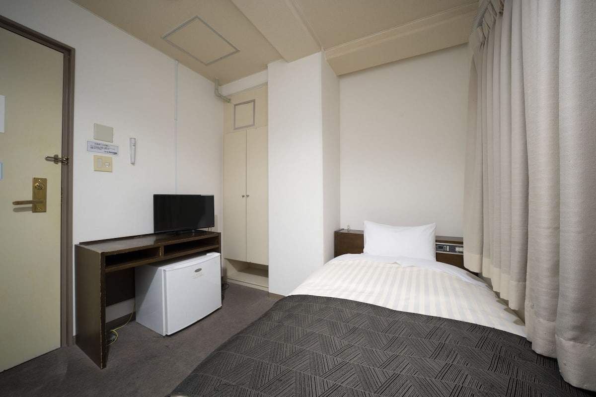 A hotel room with a bed, a desk with a television, a small refrigerator, and a door.