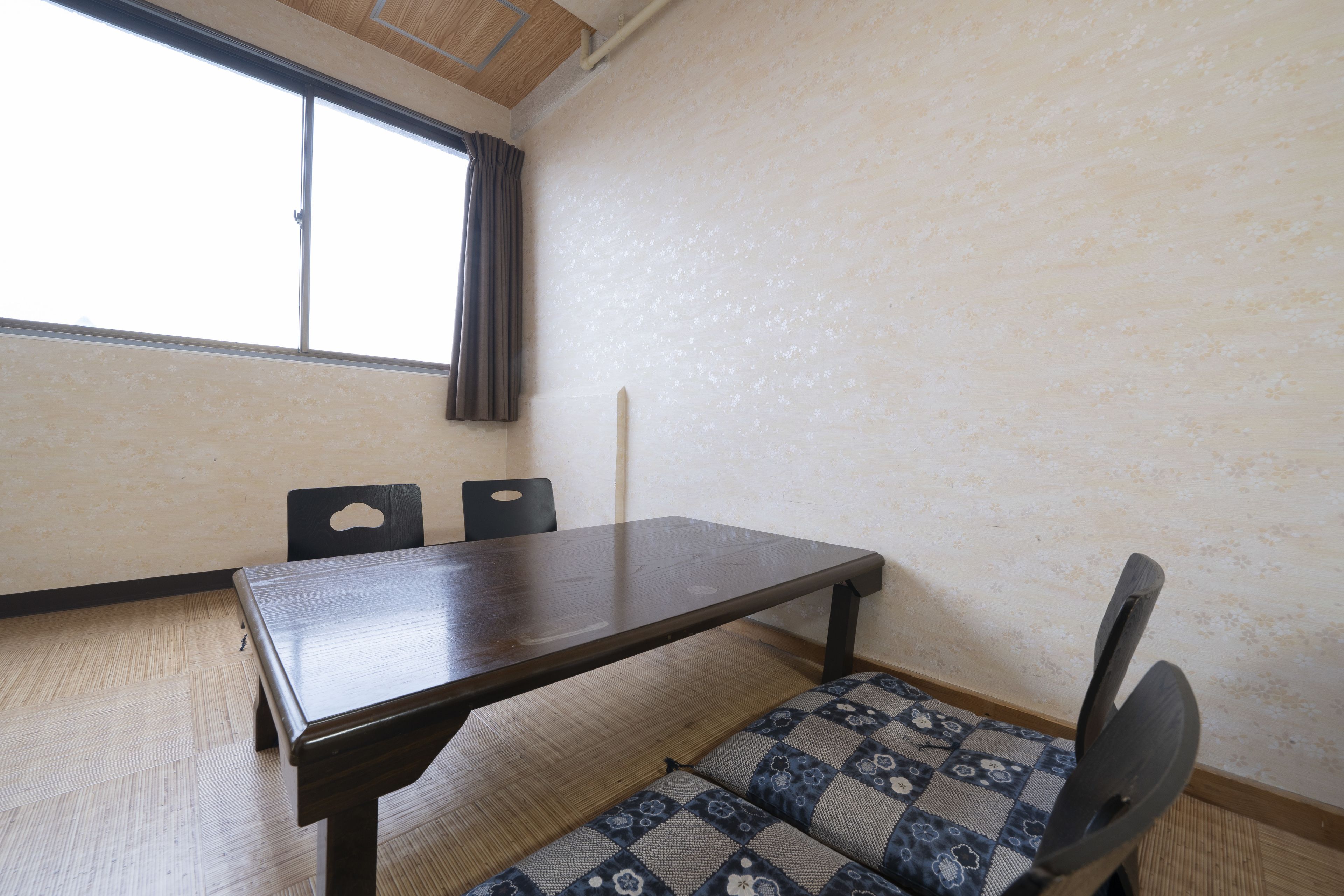 A room with a wooden table, two chairs, a patterned sofa, a window with curtains, and a textured wall.