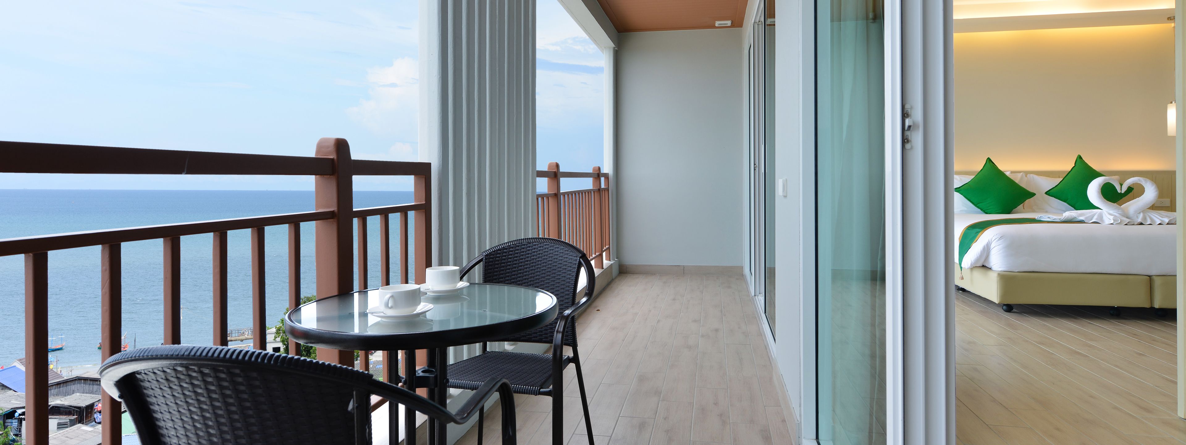 A balcony with a glass table and chairs, a bed with green pillows, and a view of the sea.