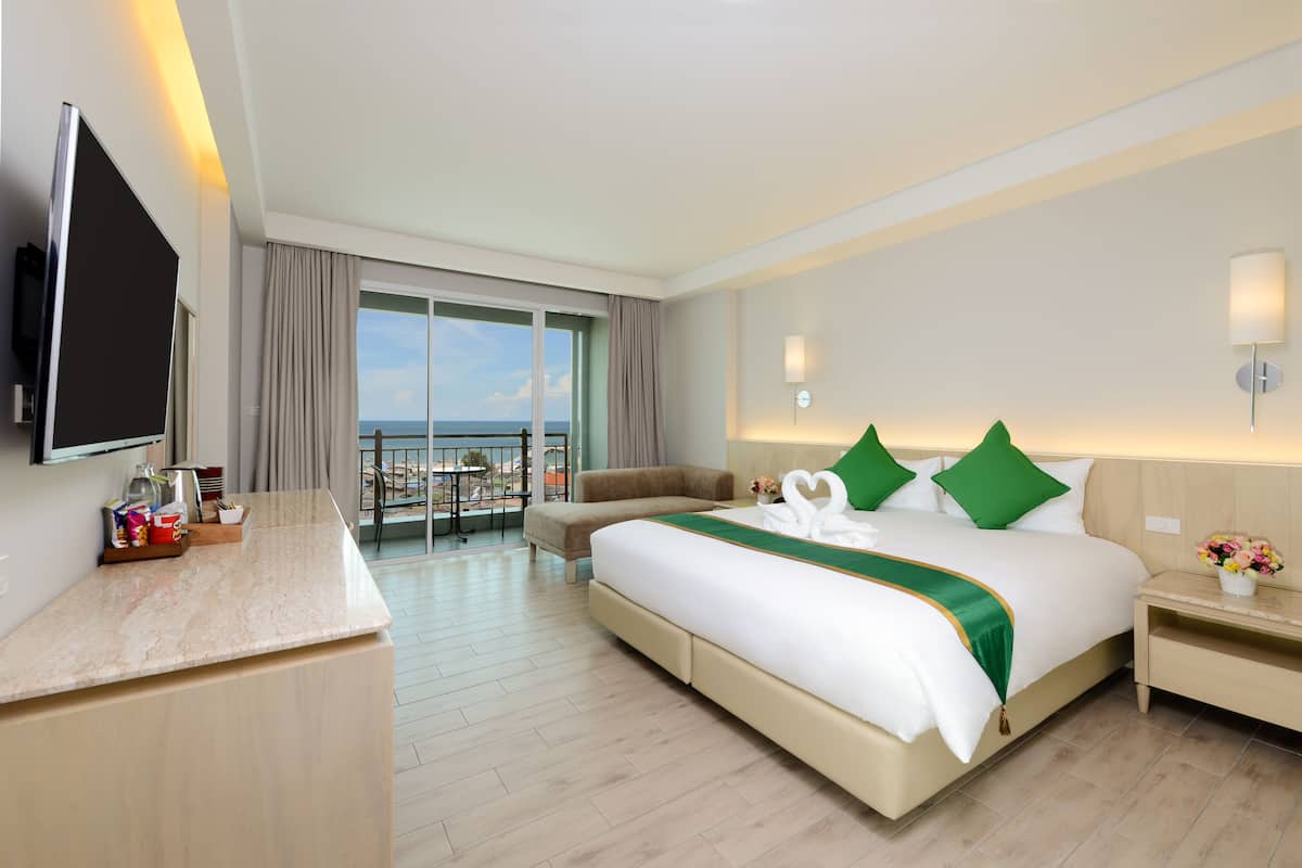 A hotel room with a bed, a desk, a TV, a balcony with a view of the sea, and a nightstand with a lamp.