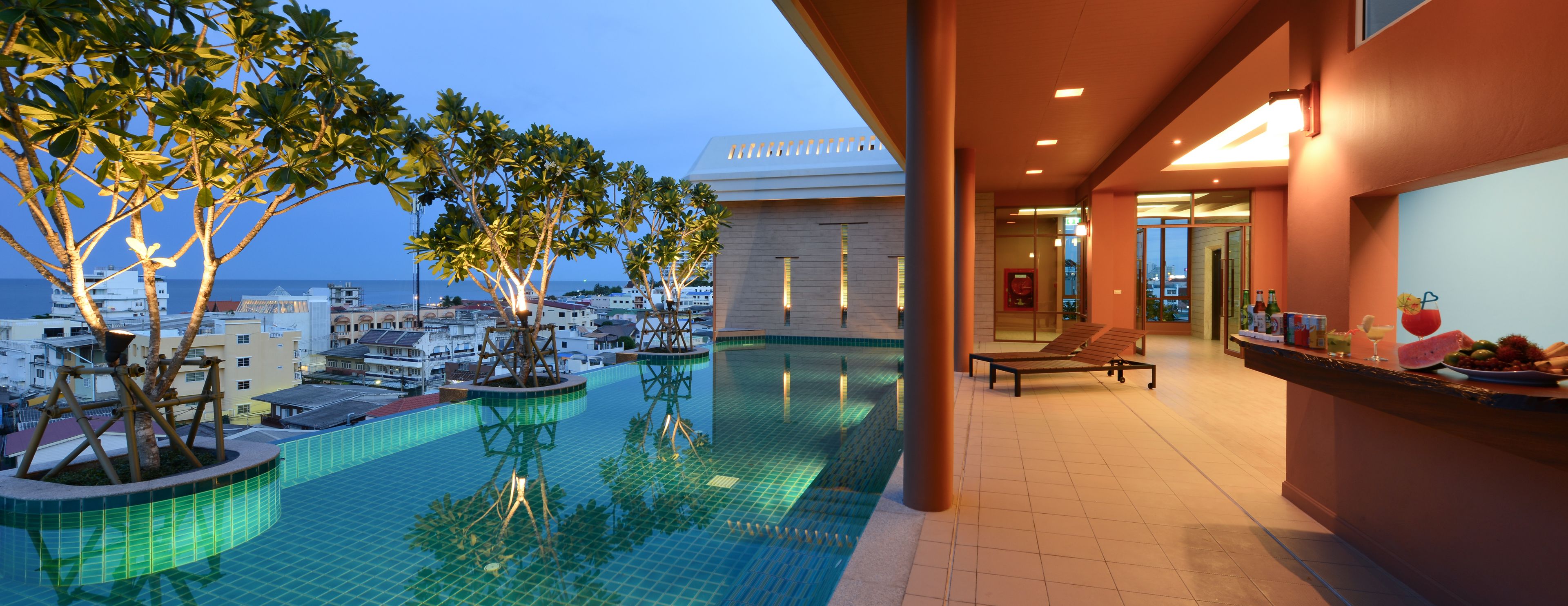 A rooftop infinity pool with a view of the cityscape, a bar area with a fruit display, and a lounge chair.