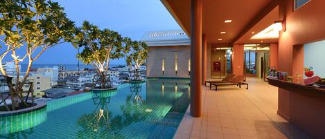 A rooftop infinity pool with a view of the cityscape, a bar area with a fruit display, and a lounge chair.