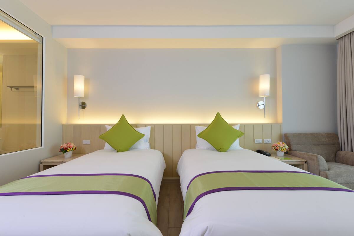 A hotel room with two beds, each with a green pillow, a nightstand with a phone and flowers, and a couch.