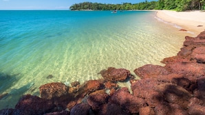 Ein Strand mit klarem türkisblauem Wasser, einem sandigen Ufer und roten Felsen im Vordergrund.