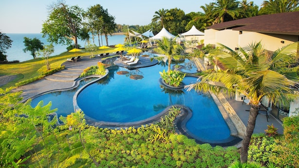 Ein Poolbereich eines Resorts mit mehreren Wasserspielen, umgeben von üppiger Vegetation und Palmen, mit Blick auf ein Gewässer.