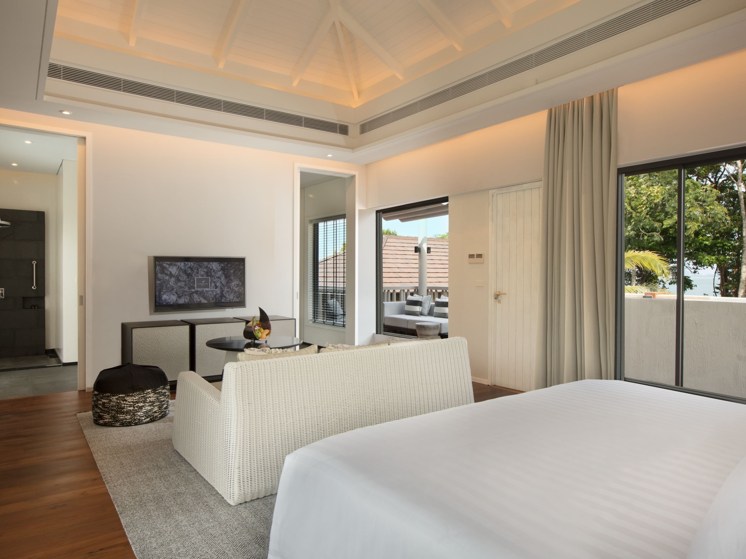 A modern hotel room with a large bed, a seating area with a white sofa and a small table, a large mirror, and a view of the outdoors through a sliding glass door.