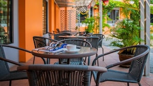 An outdoor seating area with wicker chairs and tables, some with cups and menus, surrounded by orange walls and potted plants. - Siam Boutique (Buriram)
