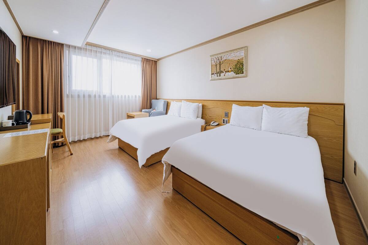 Habitación de hotel con dos camas, un escritorio con cafetera, una silla y un ventanal con cortinas.