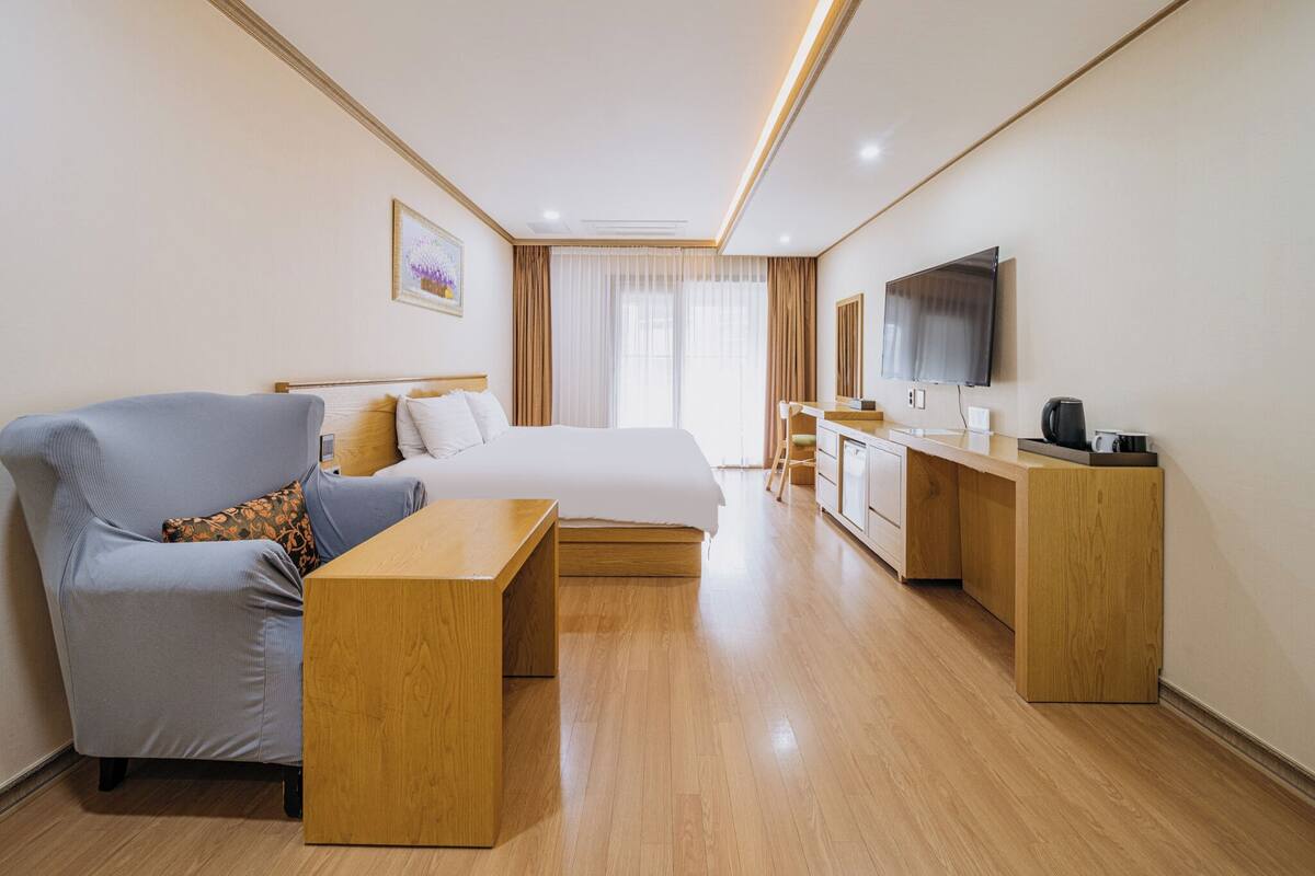 Una habitación de hotel con cama, zona de estar, escritorio, televisor y ventana con cortinas.