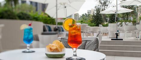 A round outdoor table with two drinks and a bowl of chips, surrounded by grey cushioned chairs and white umbrellas.