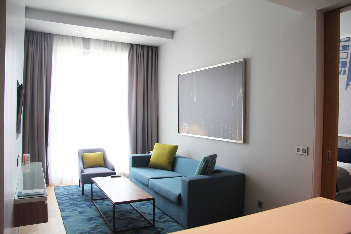 A modern hotel room with a sofa, a small table, and a large window with curtains.