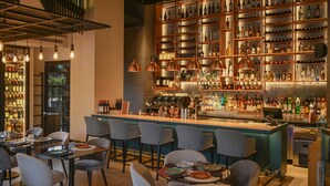 Zona de bar con varias estanterías llenas de botellas, una barra con caja registradora y varios taburetes.
