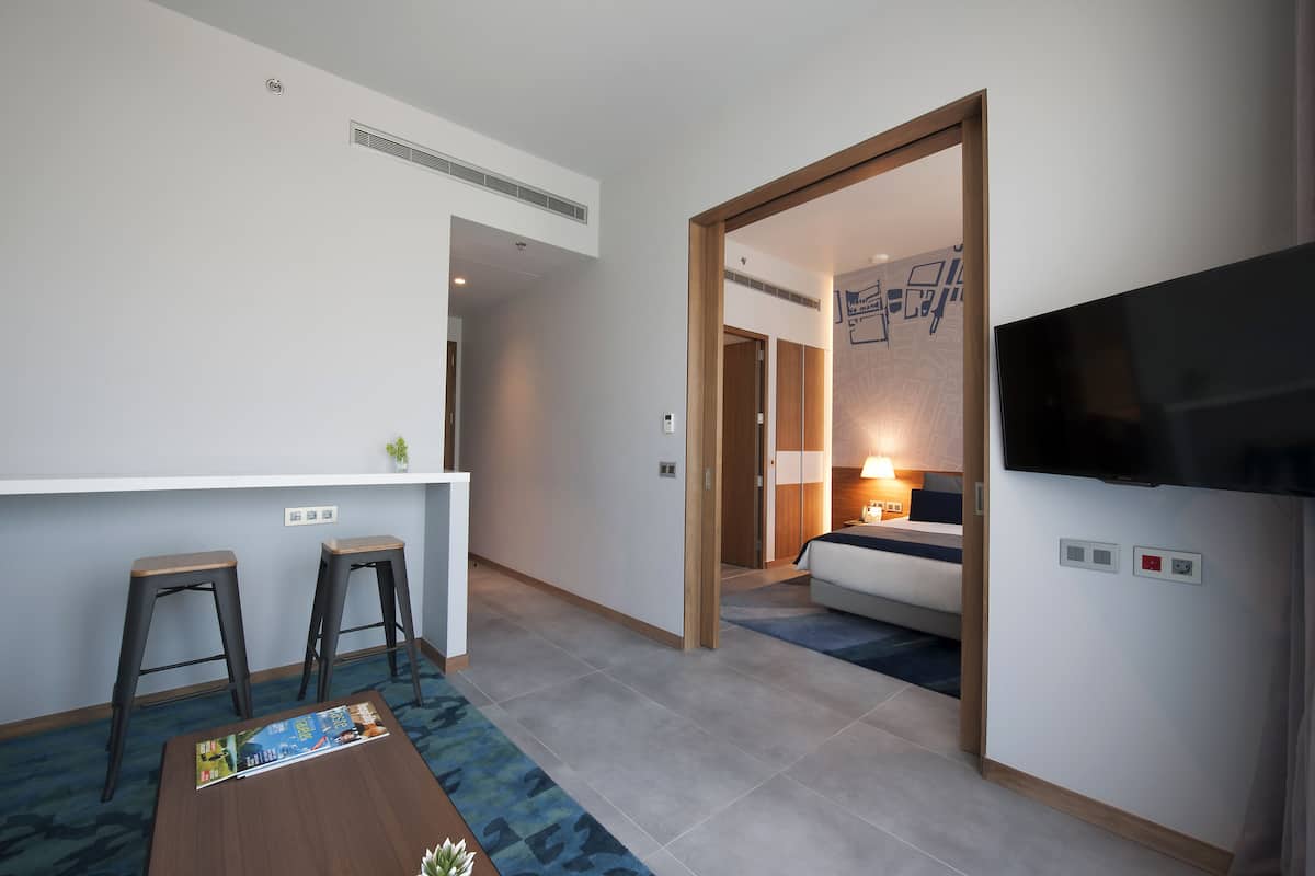 A hotel room with a TV, a bed, a desk with two stools, and a small table with a magazine.