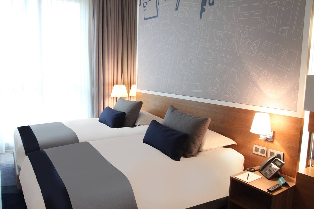 A hotel room with a large bed, two bedside tables with lamps, a wall with a cityscape mural, and a window with curtains.