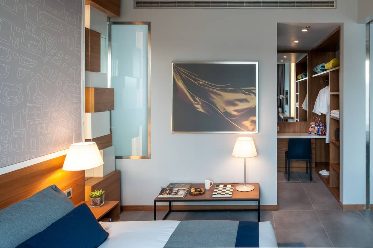A modern hotel room with a bed, bedside table, lamp, artwork, and a view into the bathroom.