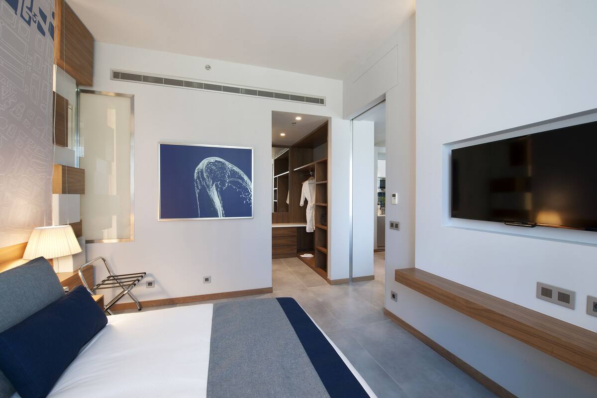 A modern hotel room with a bed, a TV mounted on the wall, a walk-in closet, and a bathroom visible through an open door.