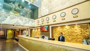 A hotel reception area with a receptionist, multiple clocks, and a large golden wall. - Nan Fang Da Sha Hotel (Guangzhou)