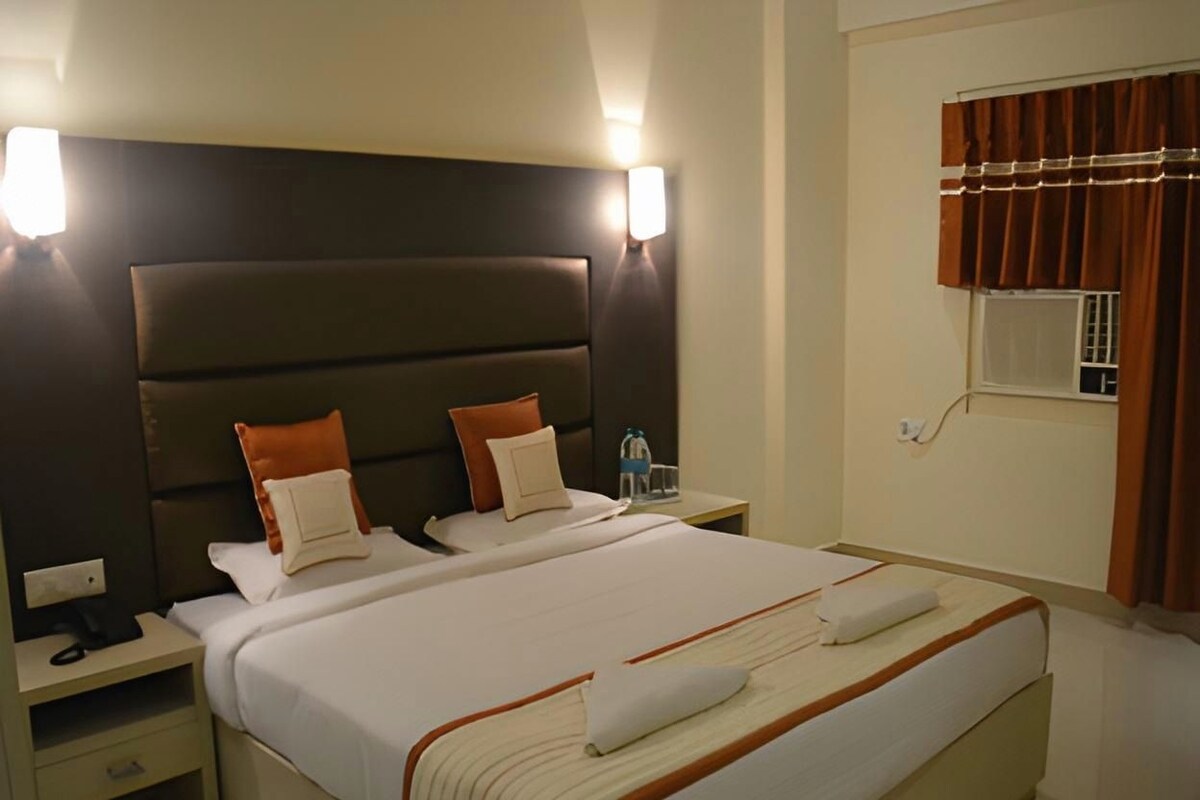 A hotel room with a large bed, two bedside tables, a nightstand, a wall-mounted lamp, and a window with curtains.