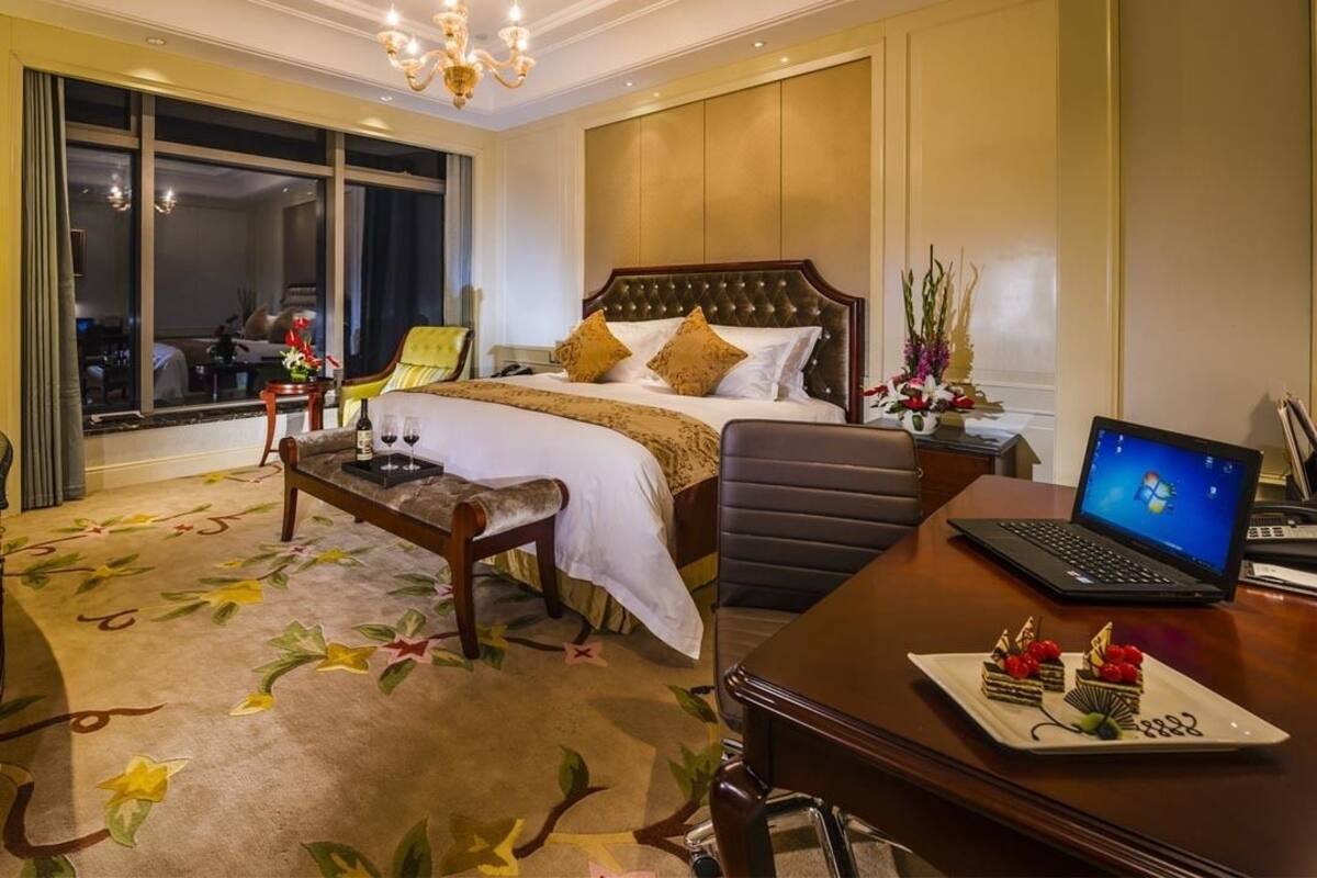 A hotel room with a large bed, a desk with a laptop, a dining table with a cake, and a view of the city through the window.