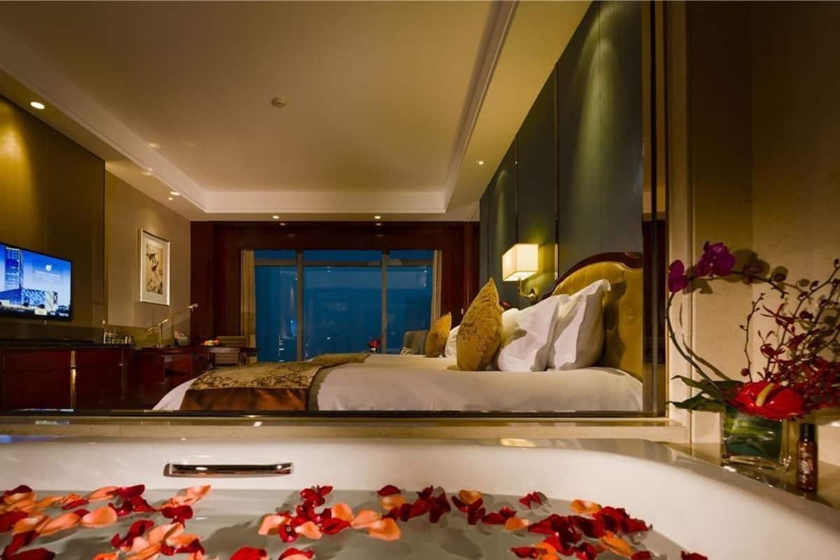 A hotel room with a bed, a television, a desk, and a bathroom with rose petals on the floor.