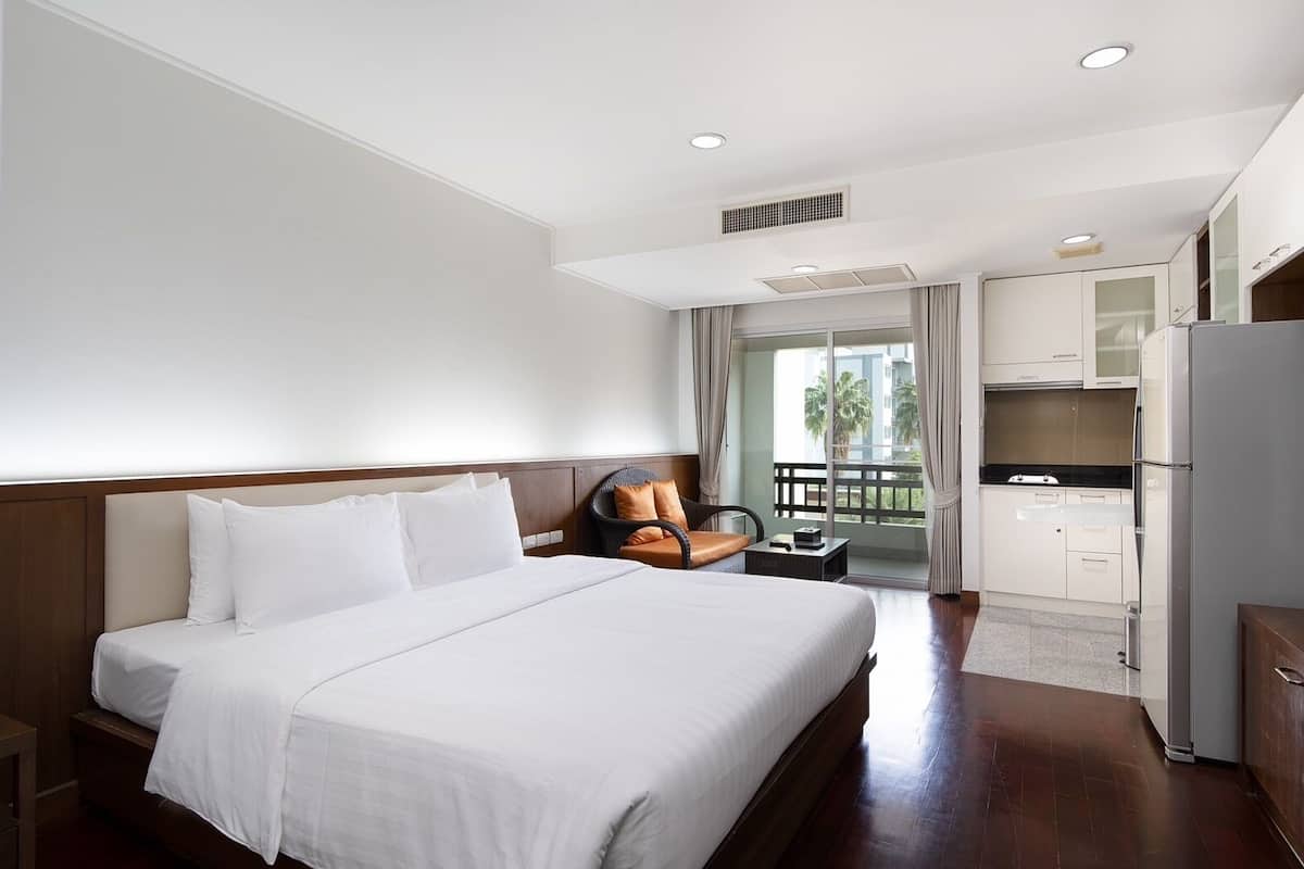 Habitación de hotel con una cama grande, un área de descanso pequeña, kitchenette y balcón con vistas a palmeras.