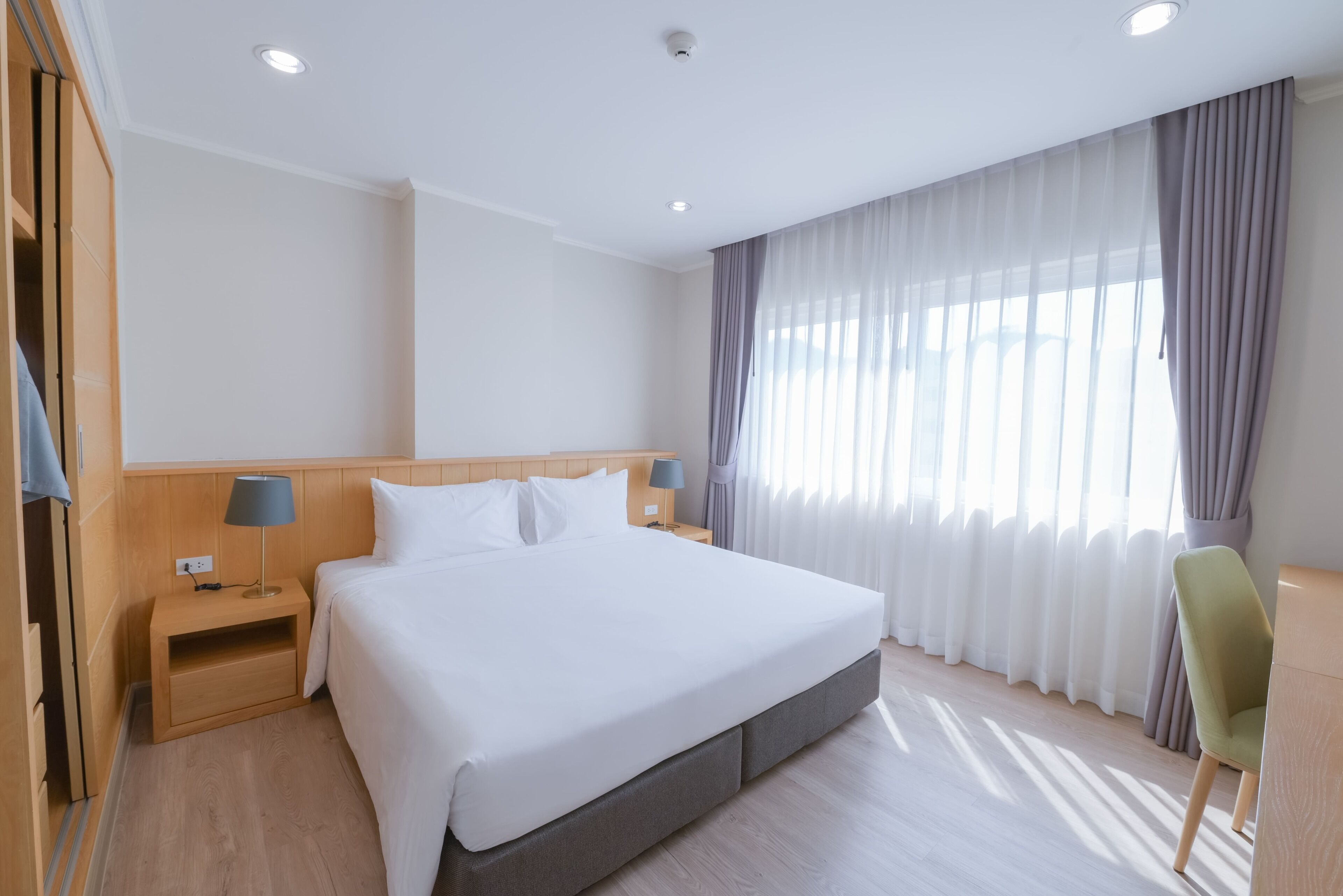 Ein Hotelzimmer mit einem großen Bett, einem Holz-Kopfteil, Nachttischen mit Lampen, einem grünen Sessel und einem Fenster mit durchsichtigen Vorhängen.