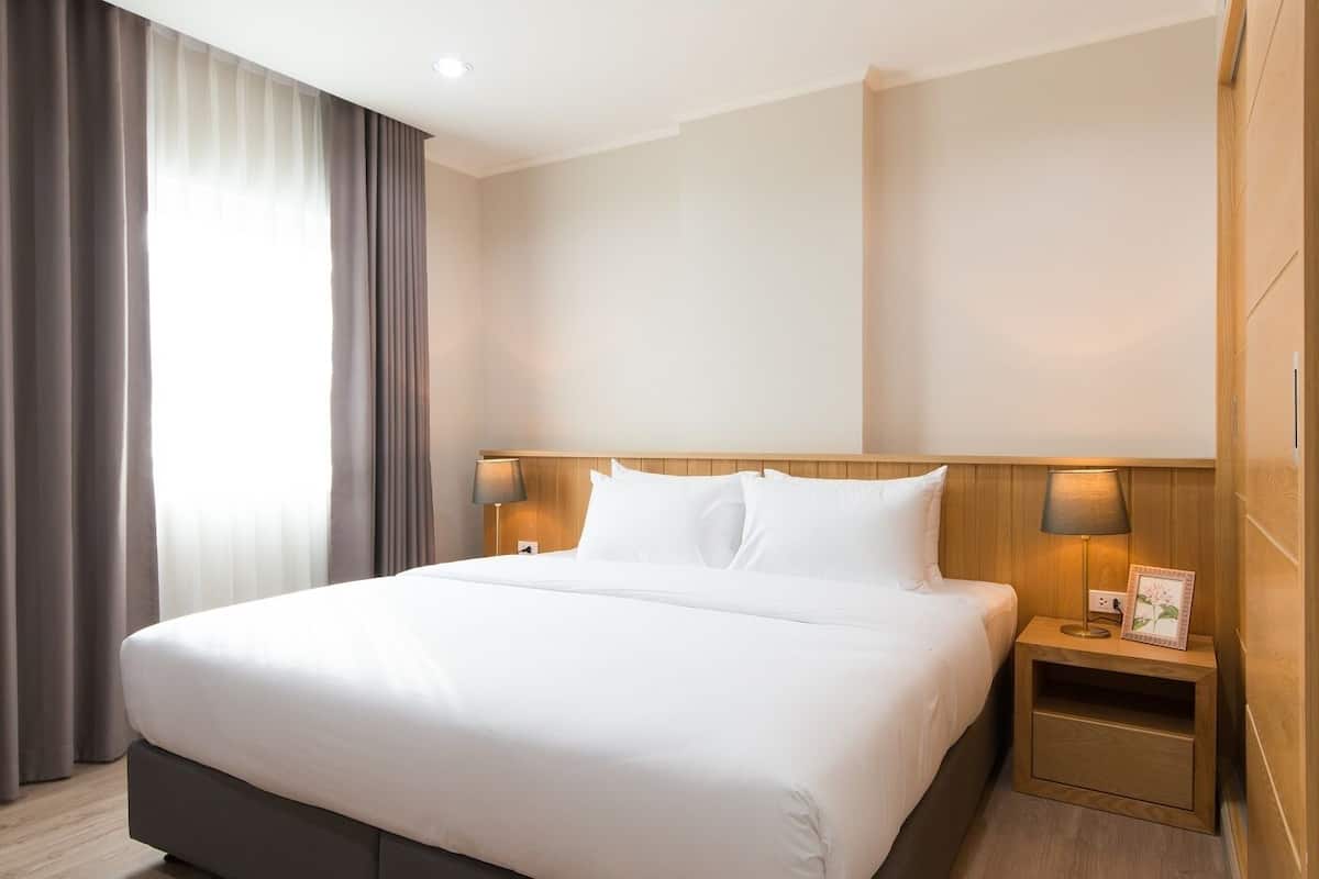 Habitación de hotel con una cama grande, cabecera de madera, dos mesitas de noche con lámparas y una ventana con cortinas.