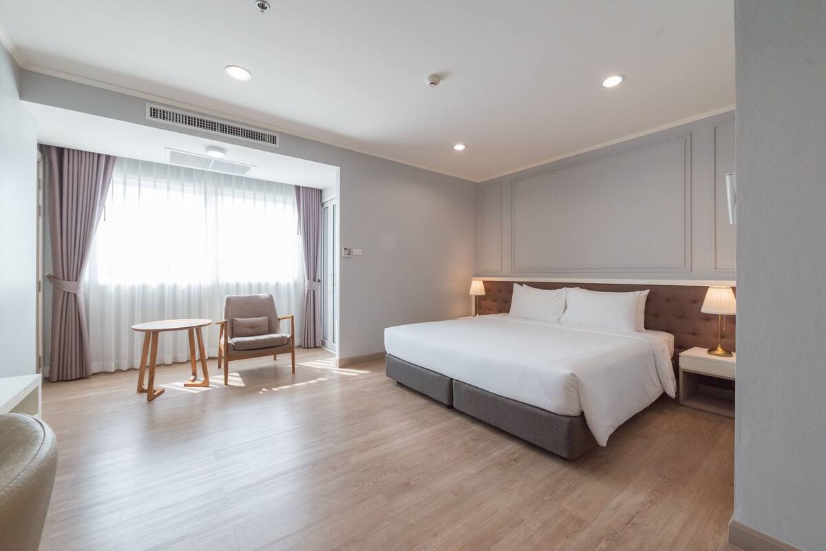A hotel room with a large bed, a bedside table with a lamp, a chair, a small table, and a window with curtains.