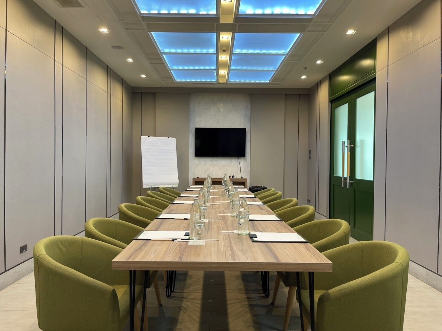 A conference room with a long table, green chairs, a whiteboard, and a flat-screen TV.