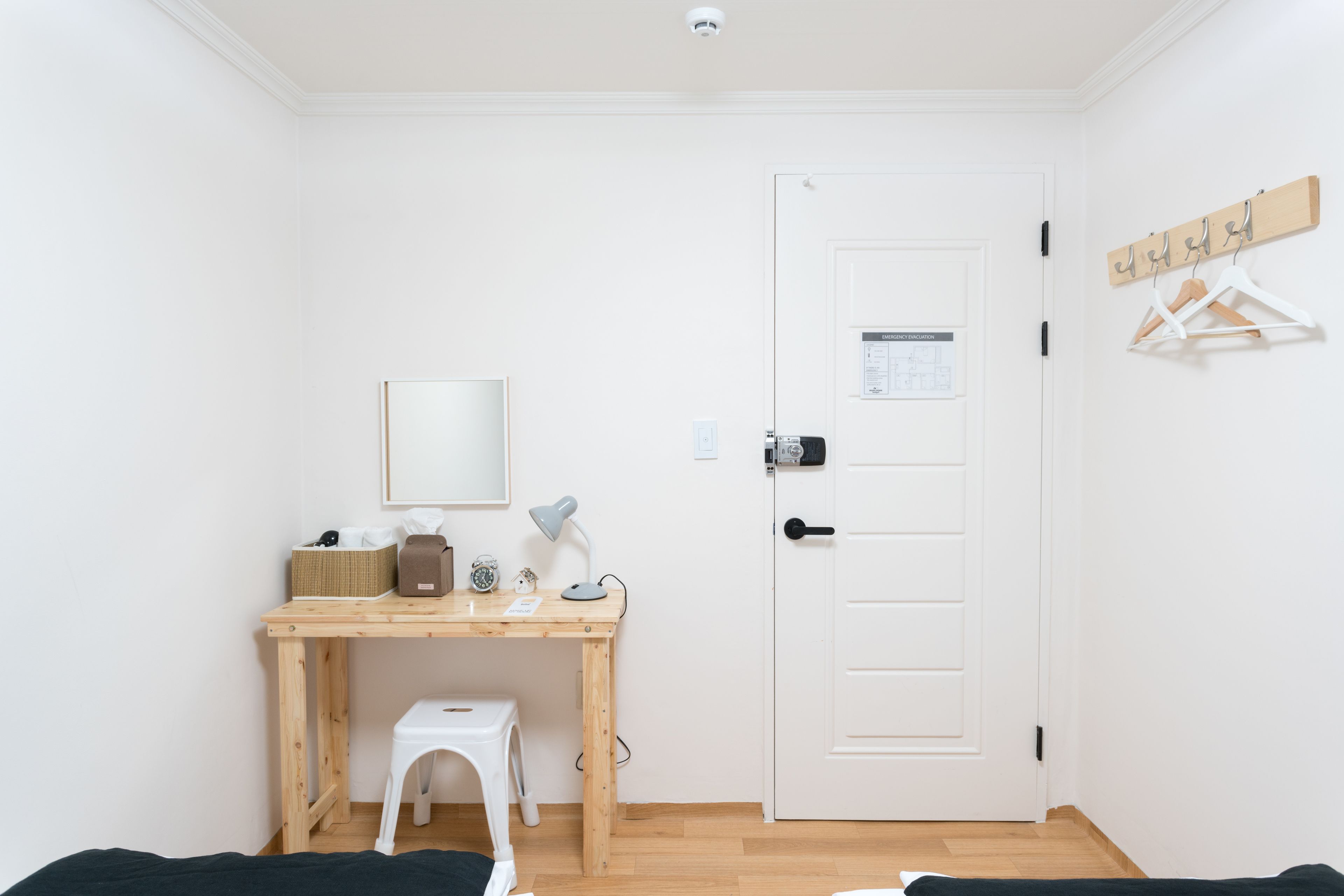 A small, clean room with a wooden desk, a white chair, a bed, and a white door with a coat rack.