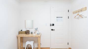 A small, clean room with a wooden desk, a white chair, a bed, and a white door with a coat rack.
