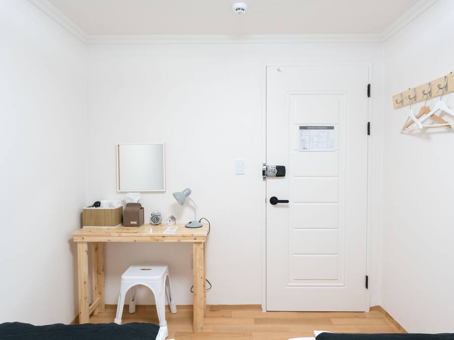 A small, clean room with a wooden desk, a white chair, a bed, and a white door with a coat rack.