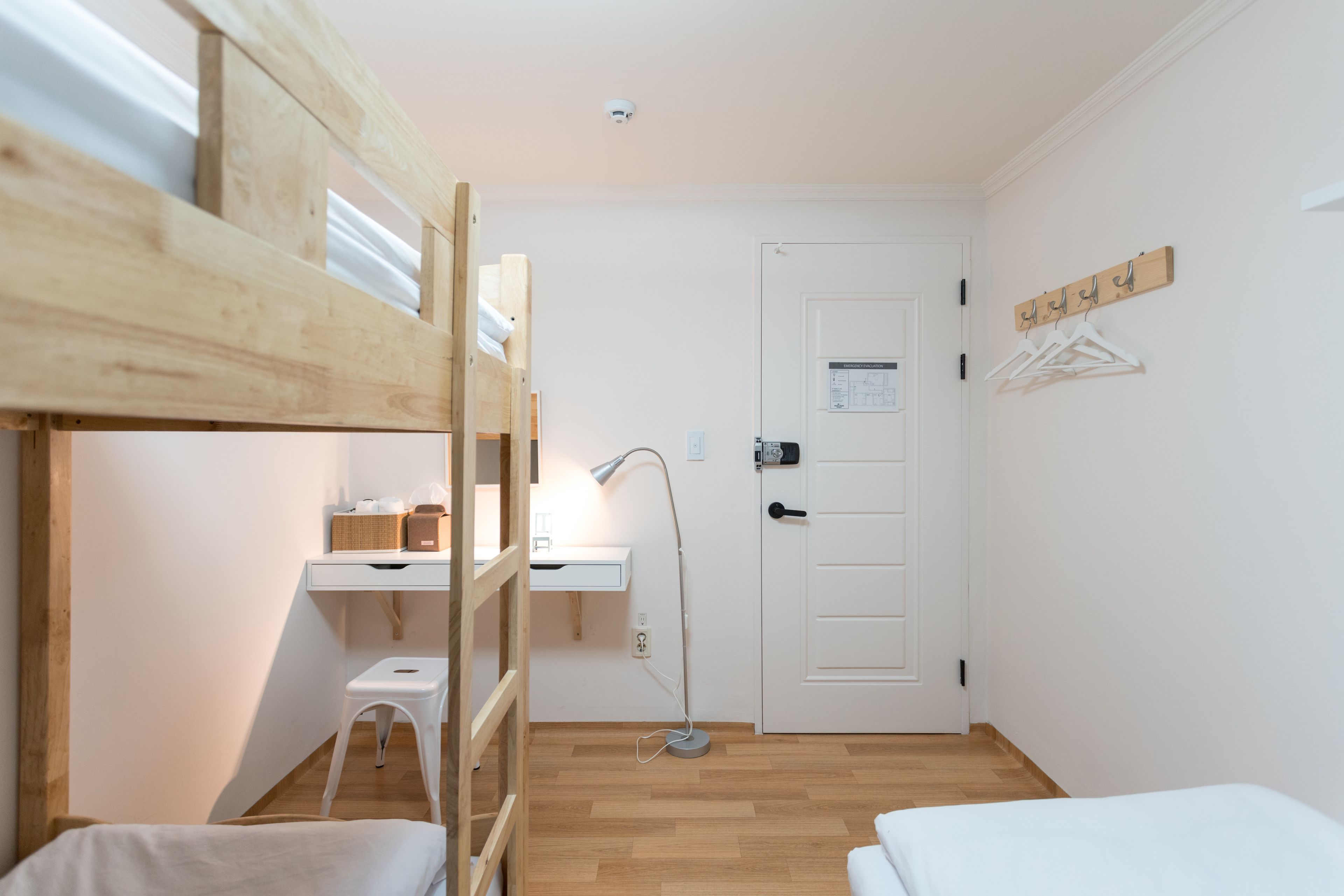 A compact room with a bunk bed, a desk with a lamp, a small stool, and a door with a coat rack.