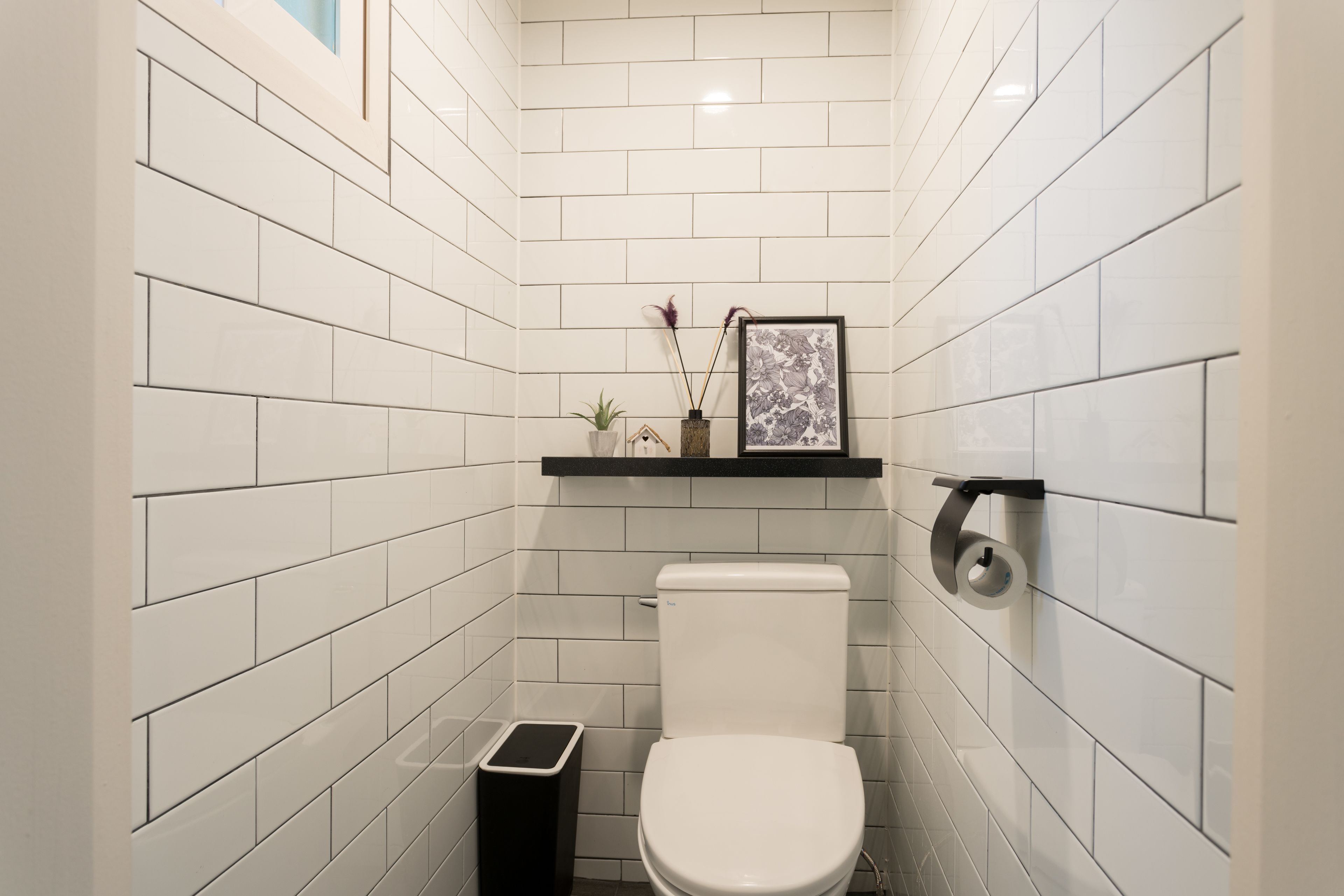 Een kleine, moderne badkamer met witte metrotegels, een wit toilet, een zwarte prullenbak, een klein plankje met een plant en een lijst, en een toiletrolhouder.
