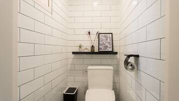 Een kleine, moderne badkamer met witte metrotegels, een wit toilet, een zwarte prullenbak, een klein plankje met een plant en een lijst, en een toiletrolhouder.