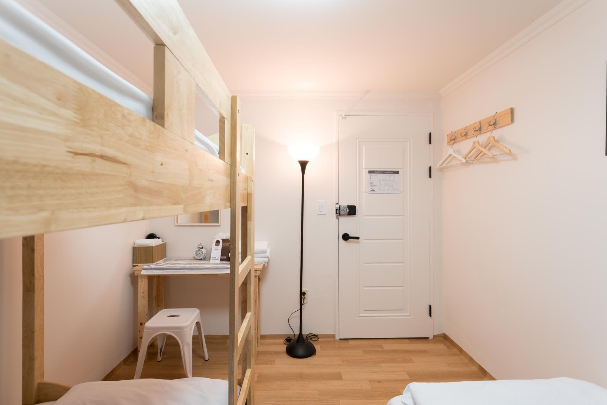 Ein kleines Zimmer mit Etagenbetten, einem Schreibtisch, einem Stuhl, einer Stehlampe und einer Tür mit Kleiderhaken.