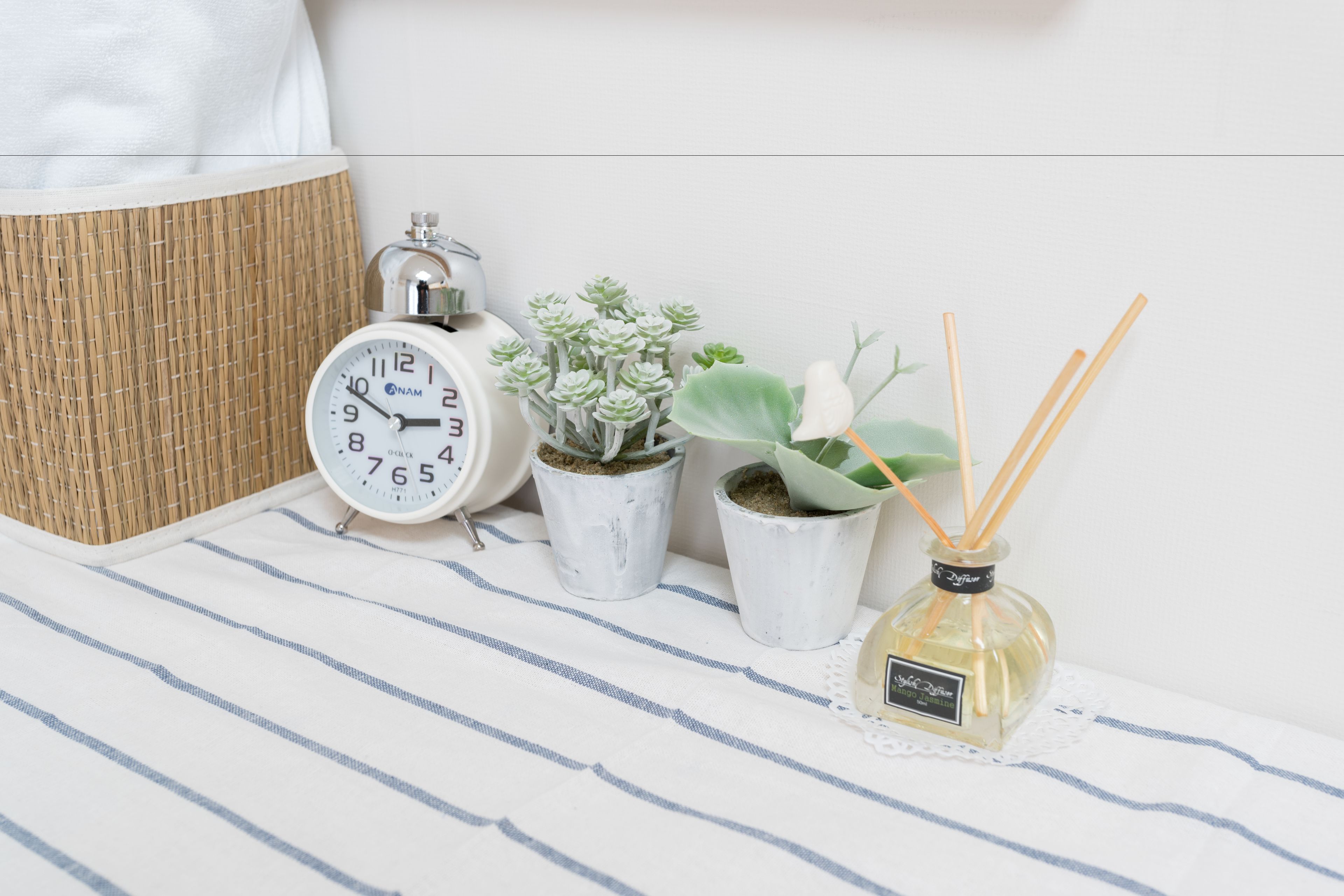 A bedside table with a striped cloth, a white analog alarm clock, two potted succulents, and a reed diffuser.