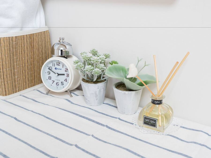 A bedside table with a striped cloth, a white analog alarm clock, two potted succulents, and a reed diffuser.