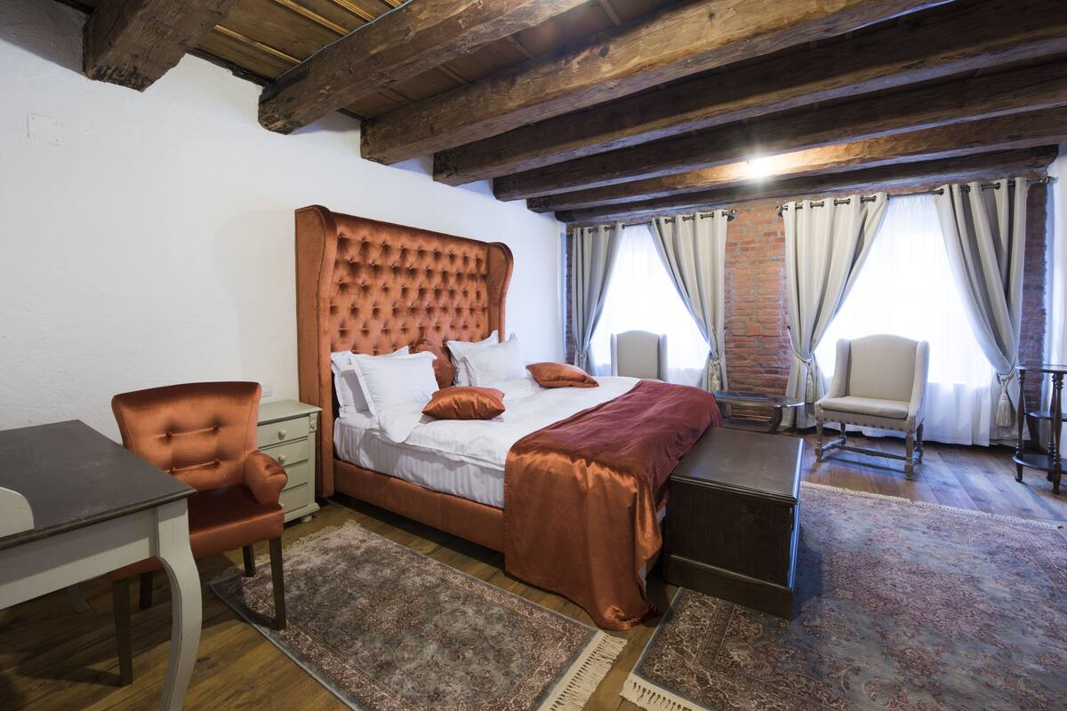 A bedroom with a large bed, a desk, and a chair. There are two windows with curtains.