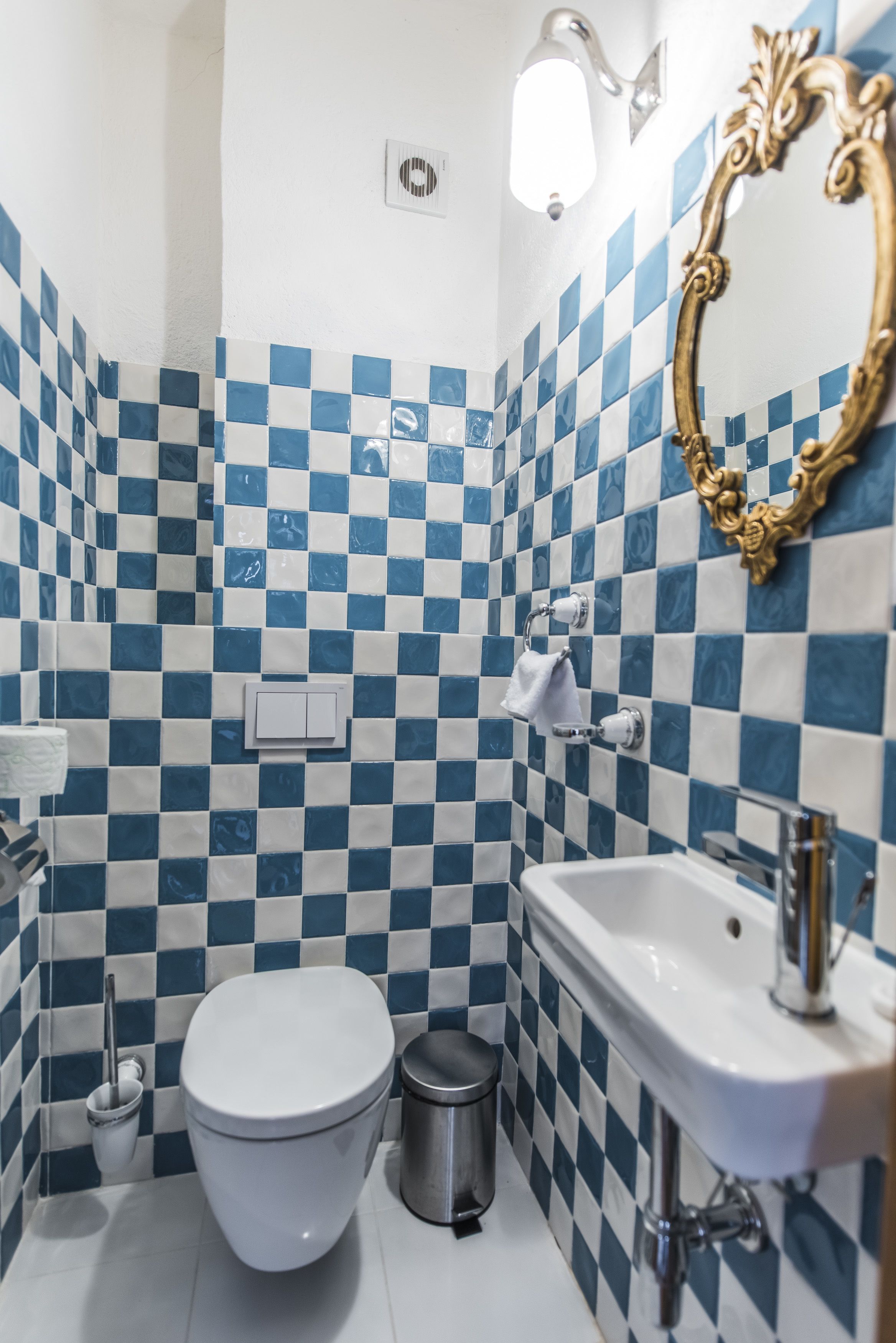 Un baño con suelo de baldosas azules y blancas en diseño de cuadros, un inodoro blanco, un lavamanos, un cubo de basura y un espejo con un marco decorado.