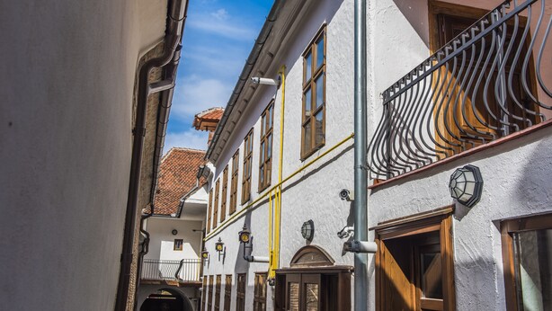 Uma rua estreita com edifícios com arquitetura tradicional, incluindo uma varanda com grades de metal ornamentadas e uma porta de madeira.